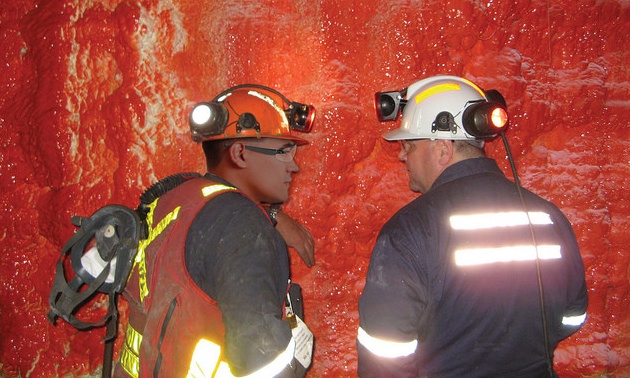 2 miners standing in an underground mine in front of a wall covered with red product. 