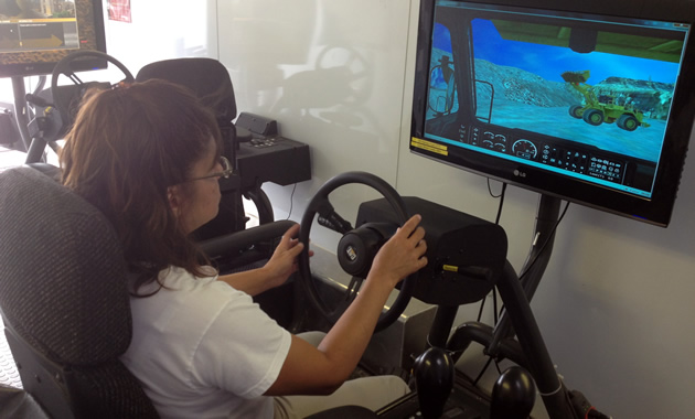 A woman sits behind a simulator steering wheel with a scenario being represented on screen.