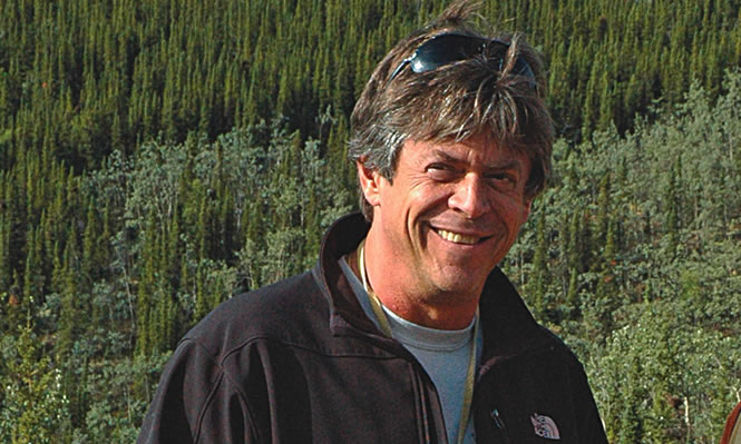 A smiling man stands against a backdrop of coniferous forest.