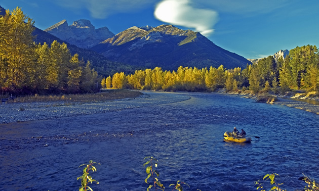 A yellow boat with fishermen floats down a deep blue river against a stunning backdrop of trees and mountains