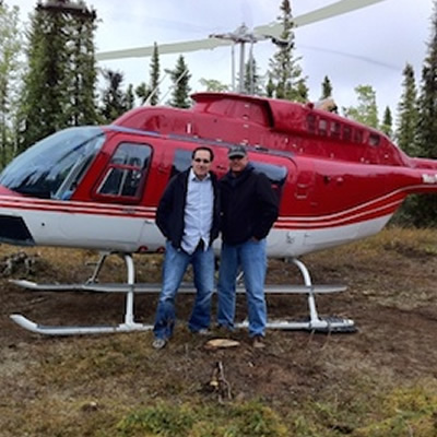 Two men standing in front of red and white helicopter in forested area. 