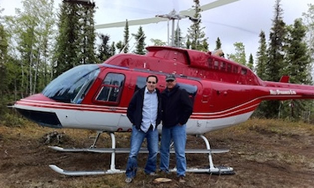 Two men standing in front of red and white helicopter in forested area. 