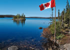 A smooth, clear blue lake with a small treed island and treed shoreline; a Canadian flag flies on the right side