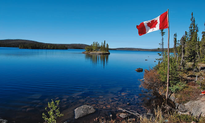 A smooth, clear blue lake with a small treed island and treed shoreline; a Canadian flag flies on the right side