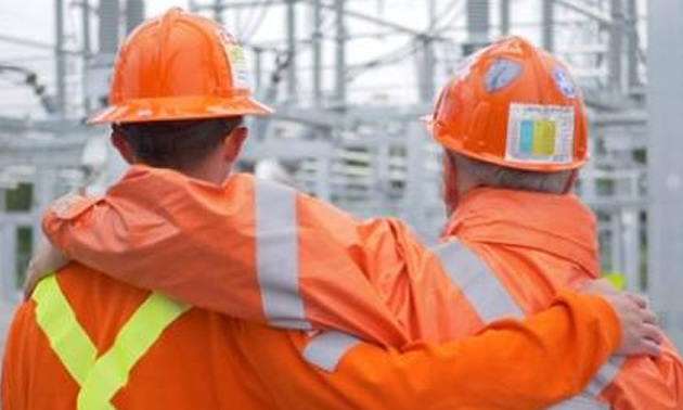 Two workers wearing orange hard hats and overalls with arms around each other's shoulders. 