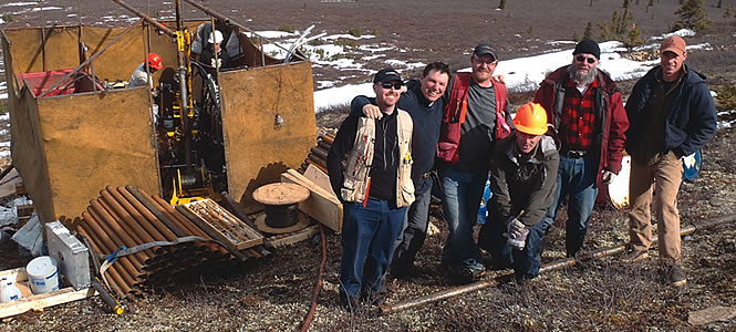 A group of six men in prospecting gear stand next to a large piece of equipment