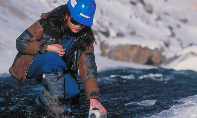 A woman, wearing a hardhat, takes a water sample from a stream.