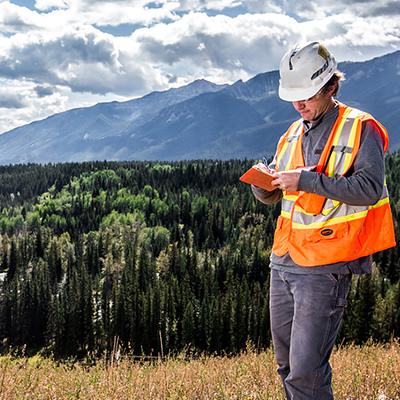 Picture of man wearing hi-vis vest and hardhat, standing on hill with mountains in background. 
