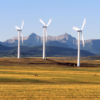 Three wind turbines are located on flat land near an irrigated field with mountains in the background.